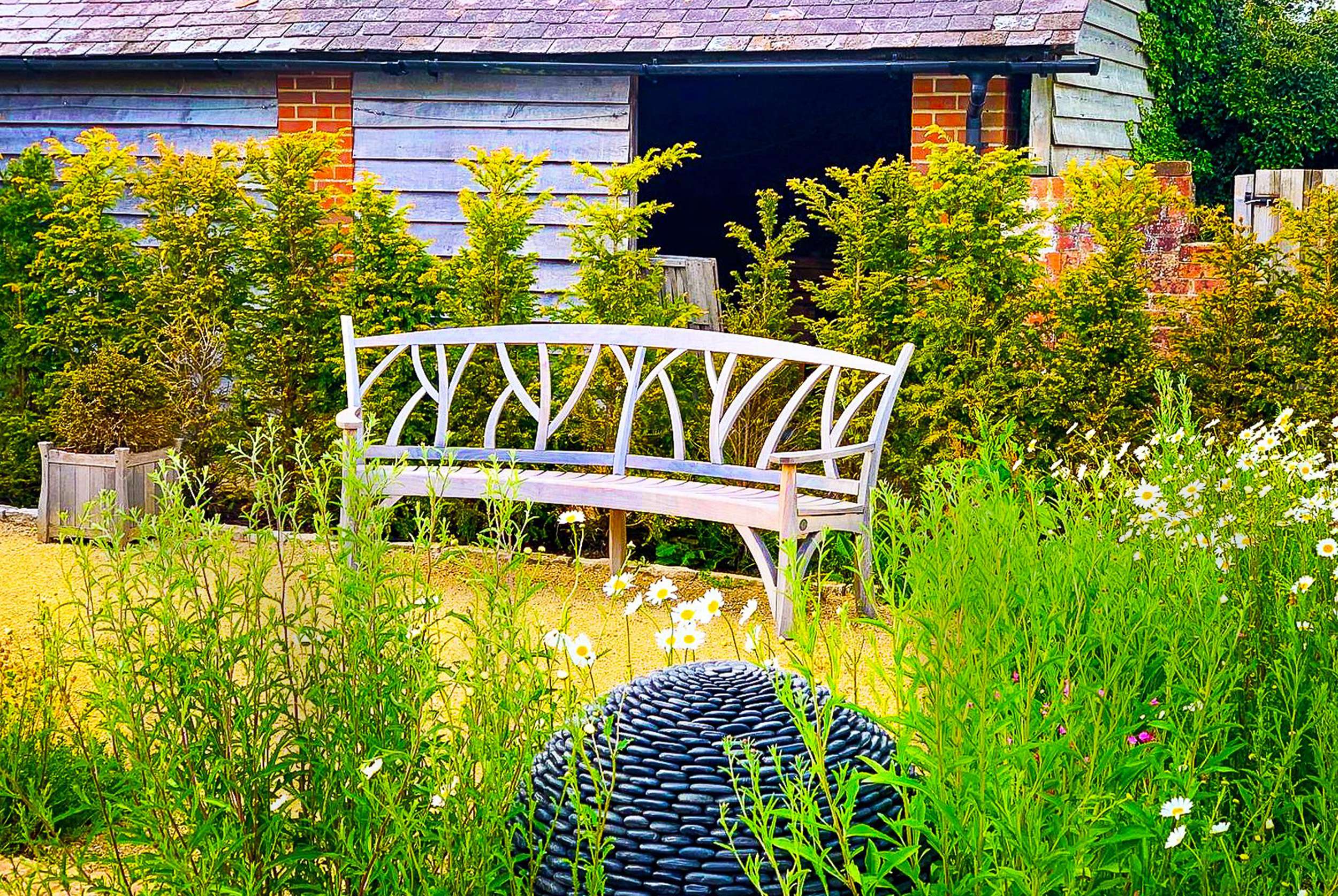 Gaze Burvill wooden bench sits in the centre with a conifer hedge behind. Cottage style planting design cross the foreground of the image with a spherical sculpture covered in pebbles sitting nestled in the planting. Designed by Hampshire based garden design company, Ann-Marie Powell Gardens.